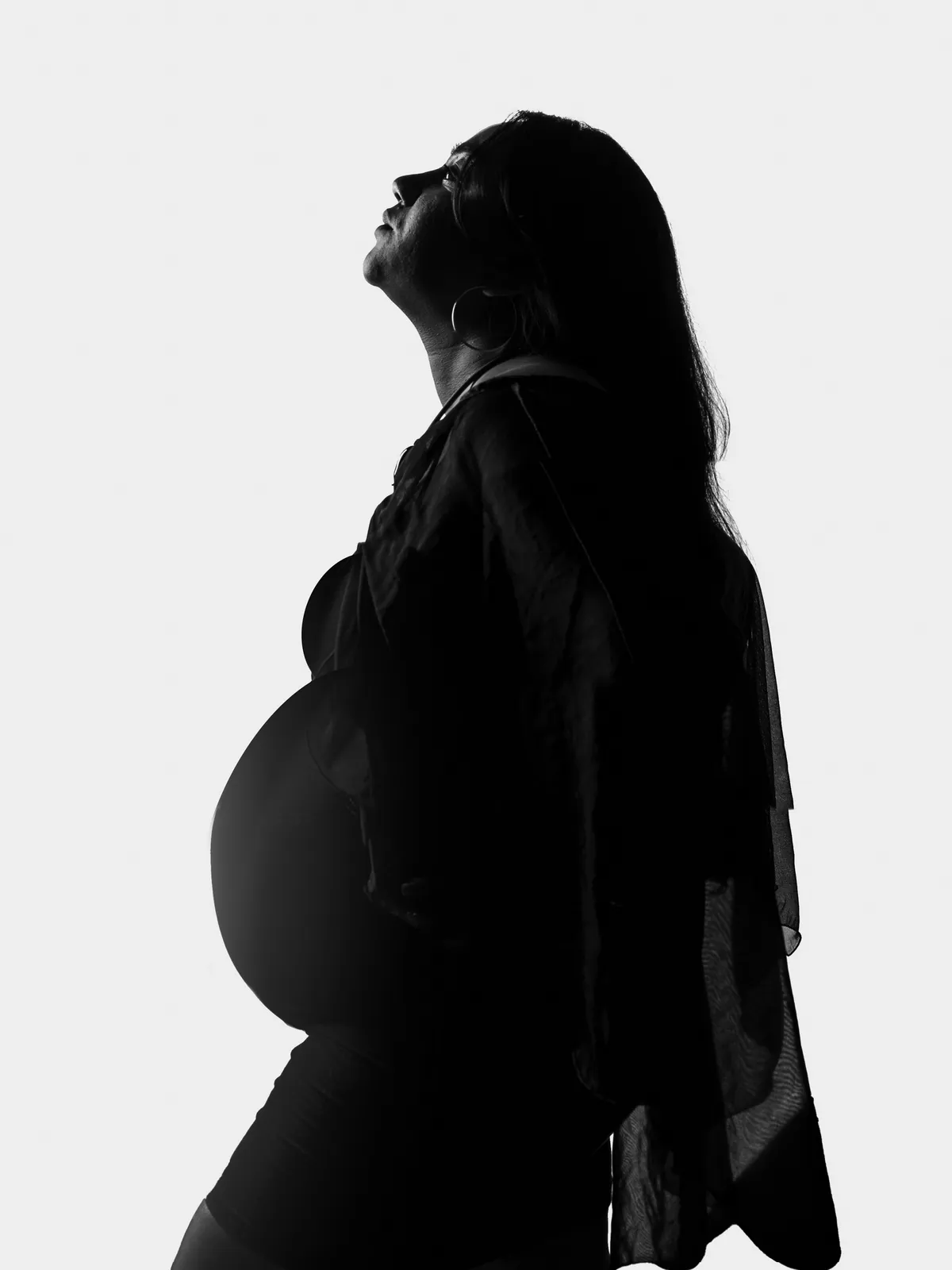 Black-and-white silhouette portrait of a pregnant woman photographed in profile against a bright studio background