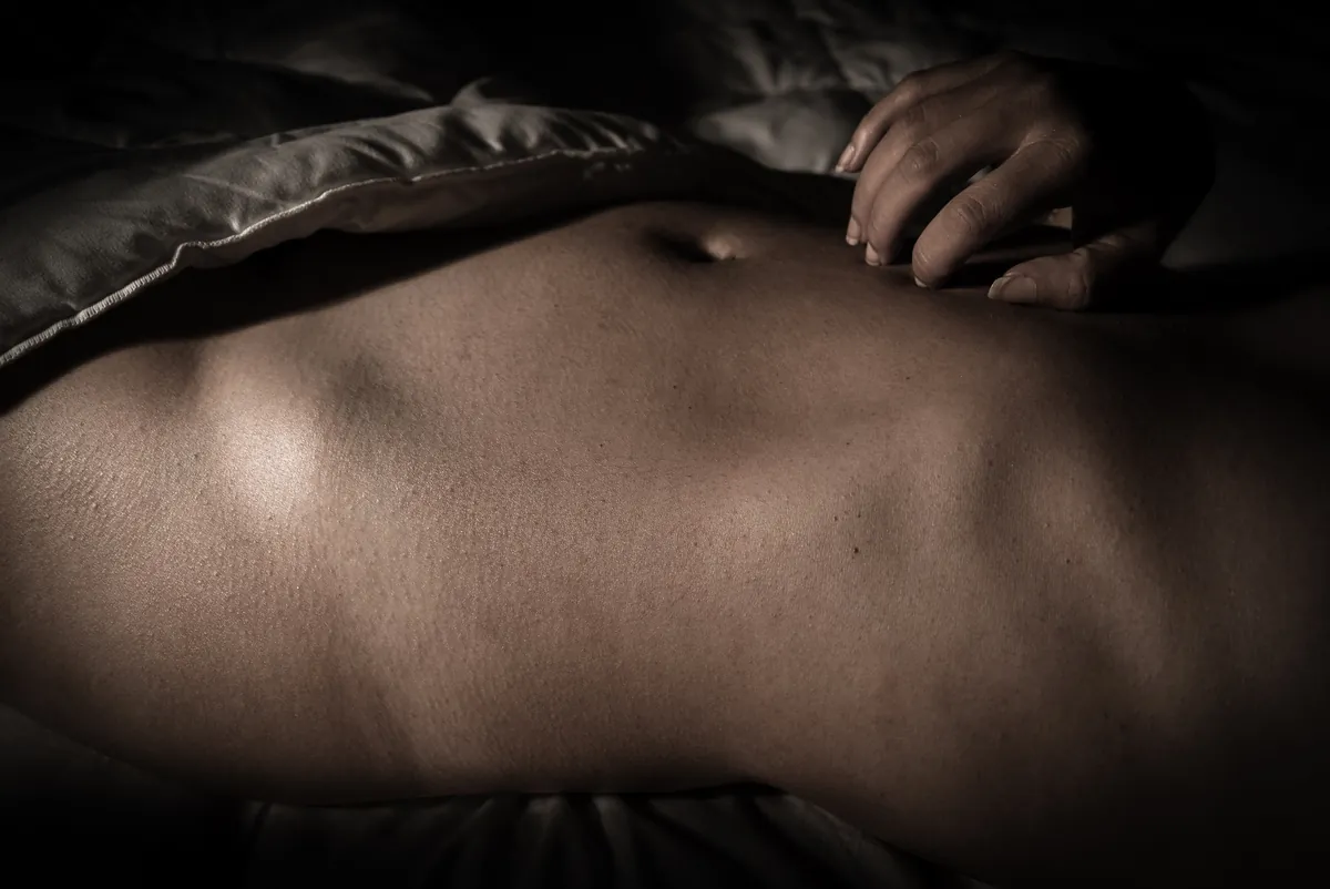 Close-up fine-art nude study of a woman's torso with restrained lighting and hand placement
