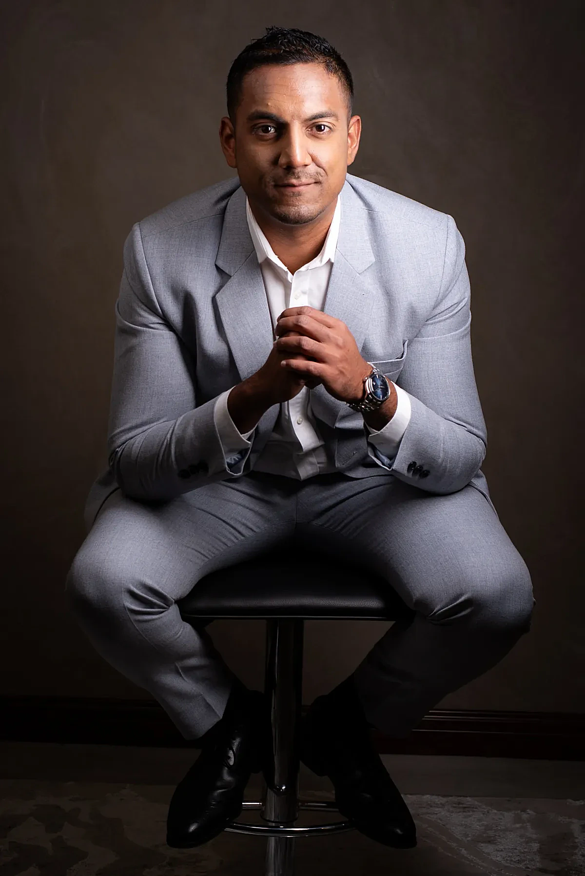 Full-body professional portrait of Sharma Soneechora seated in a grey suit