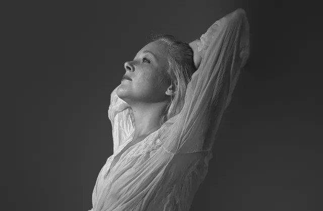 Blond girl in black panties and white top kneeling on bed in bedroom photoshoot