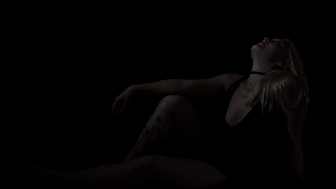 Boudoir portrait of a woman seated against a dark background, looking upward, legs extended in silhouette.